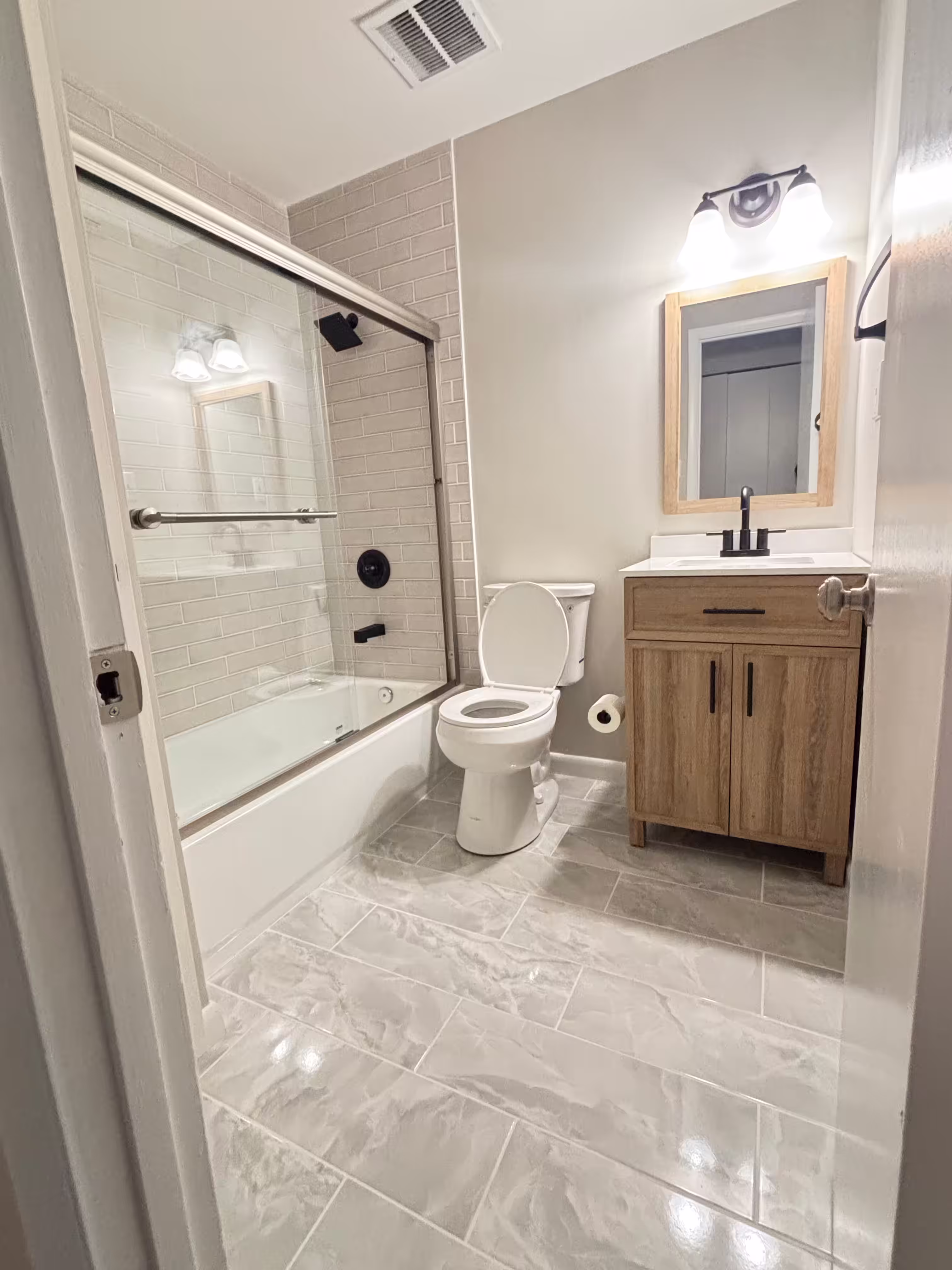 Renovated bathroom with a modern design featuring a white toilet, wood vanity with a black faucet, and a matching framed mirror. The shower has beige subway tile walls, black fixtures, and sliding glass doors. The floor is updated with large, glossy marble-look tiles, and the walls are painted a light neutral color. Stylish lighting fixtures are mounted above the mirror.