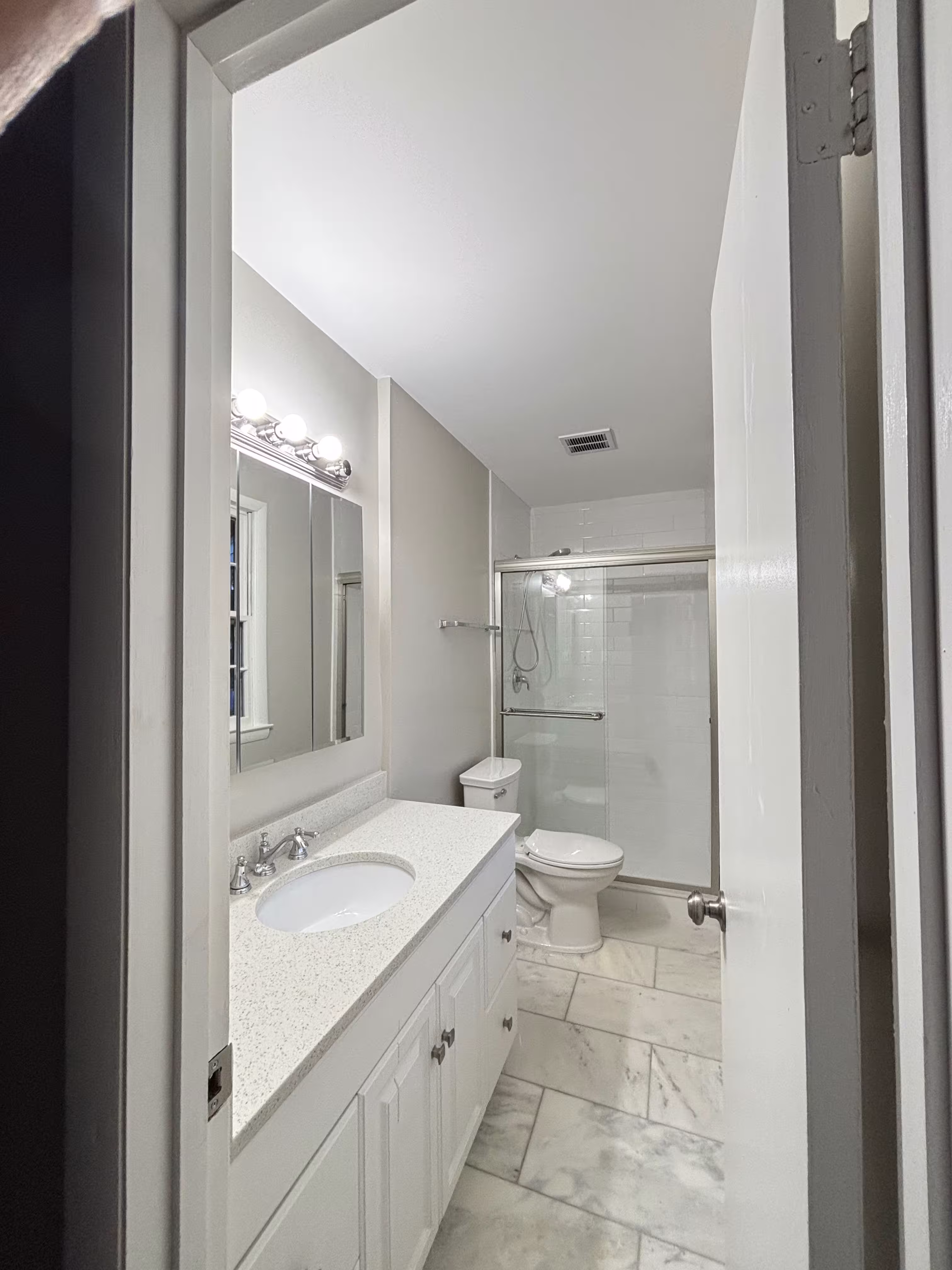 A recently renovated bathroom featuring light gray walls and a light-colored marble-patterned tile floor laid in a rectangular pattern. A white vanity with a light speckled countertop holds a white oval sink and a chrome faucet. Above the vanity is a wide, framed mirror with three multi-bulb light fixtures above it. To the right of the vanity is a white toilet, and beyond that is a shower enclosed by clear glass doors with silver hardware. A white door is partially open on the right side of the frame, showing the entrance to the bathroom. The overall impression is clean and modern.