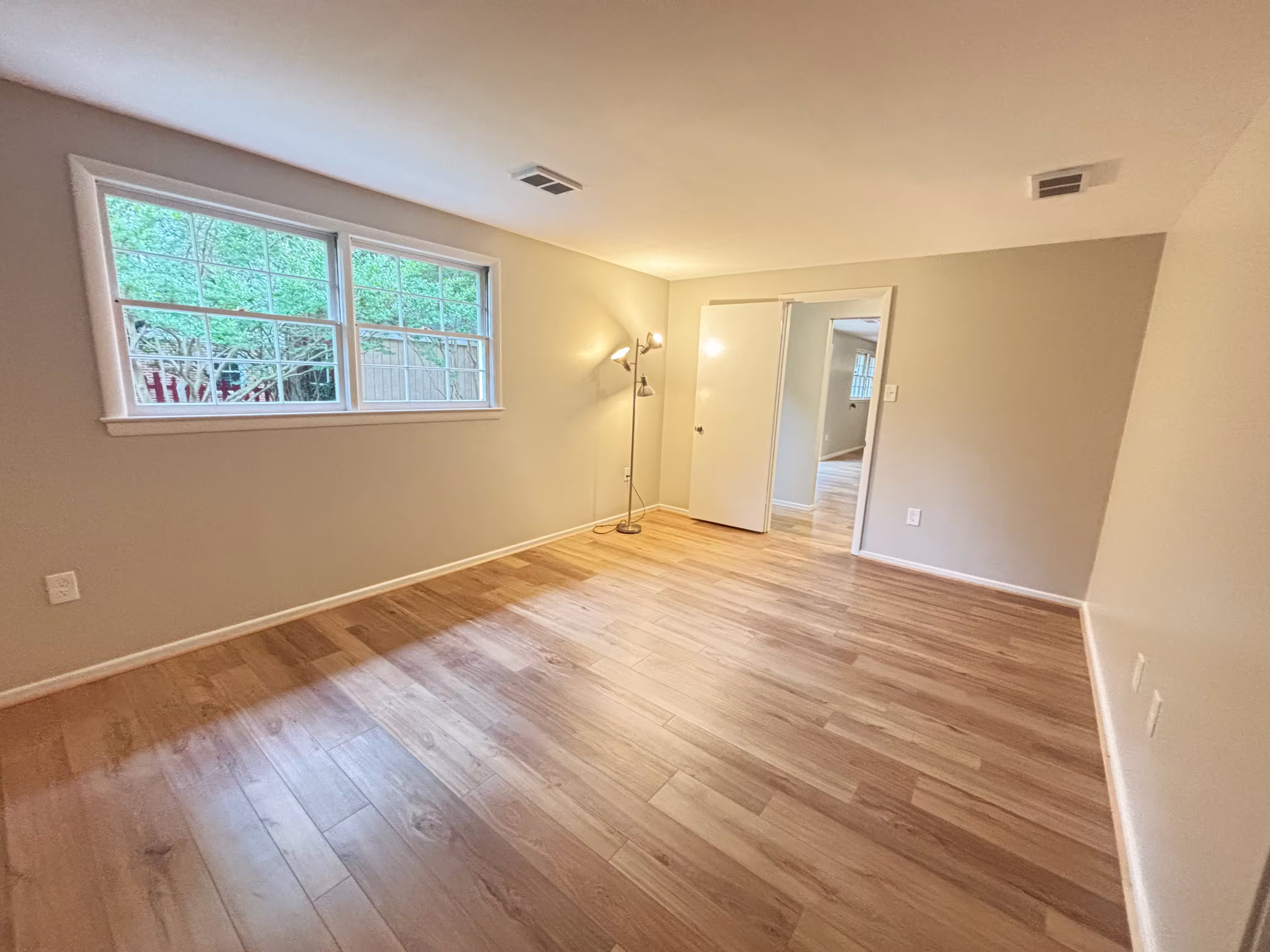 A bright and airy bedroom with light beige walls and newly installed light-colored hardwood flooring laid diagonally. A large window on the left wall looks out onto a green, leafy outdoor scene. A modern floor lamp with multiple adjustable lights stands in the corner near the window. An open white door on the far wall leads to another room with lighter-colored walls and flooring. The room is empty and appears freshly renovated.
