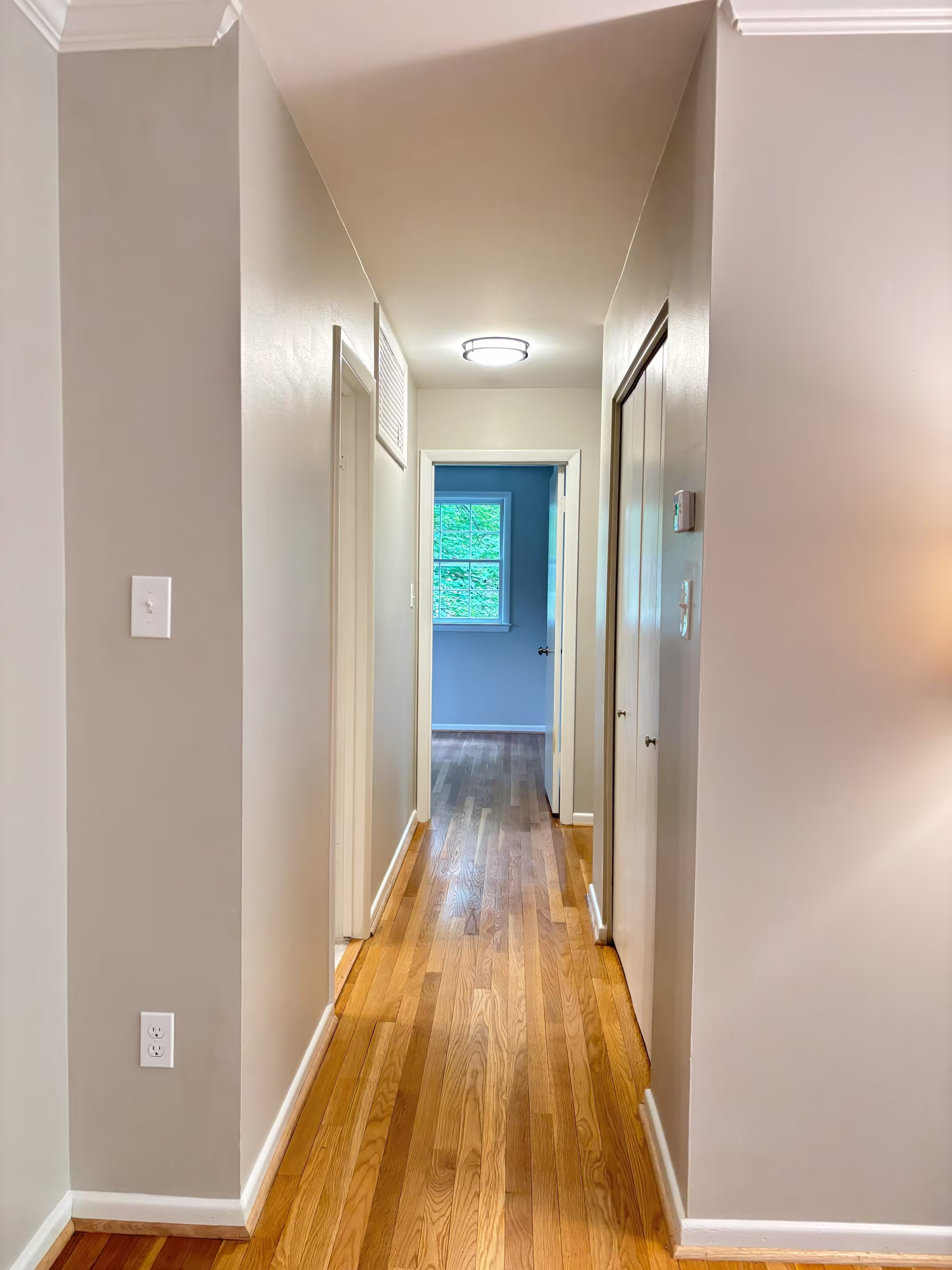 A well-lit hallway with light beige walls, white trim, and newly refinished light-colored hardwood flooring. A modern circular ceiling light illuminates the space. On the right, there's a closed sliding closet door. The hallway extends straight ahead, leading to a room with light blue walls and a window showing a green outdoor view. Another doorway is visible on the left side of the hallway. The floor has a smooth, glossy finish.
