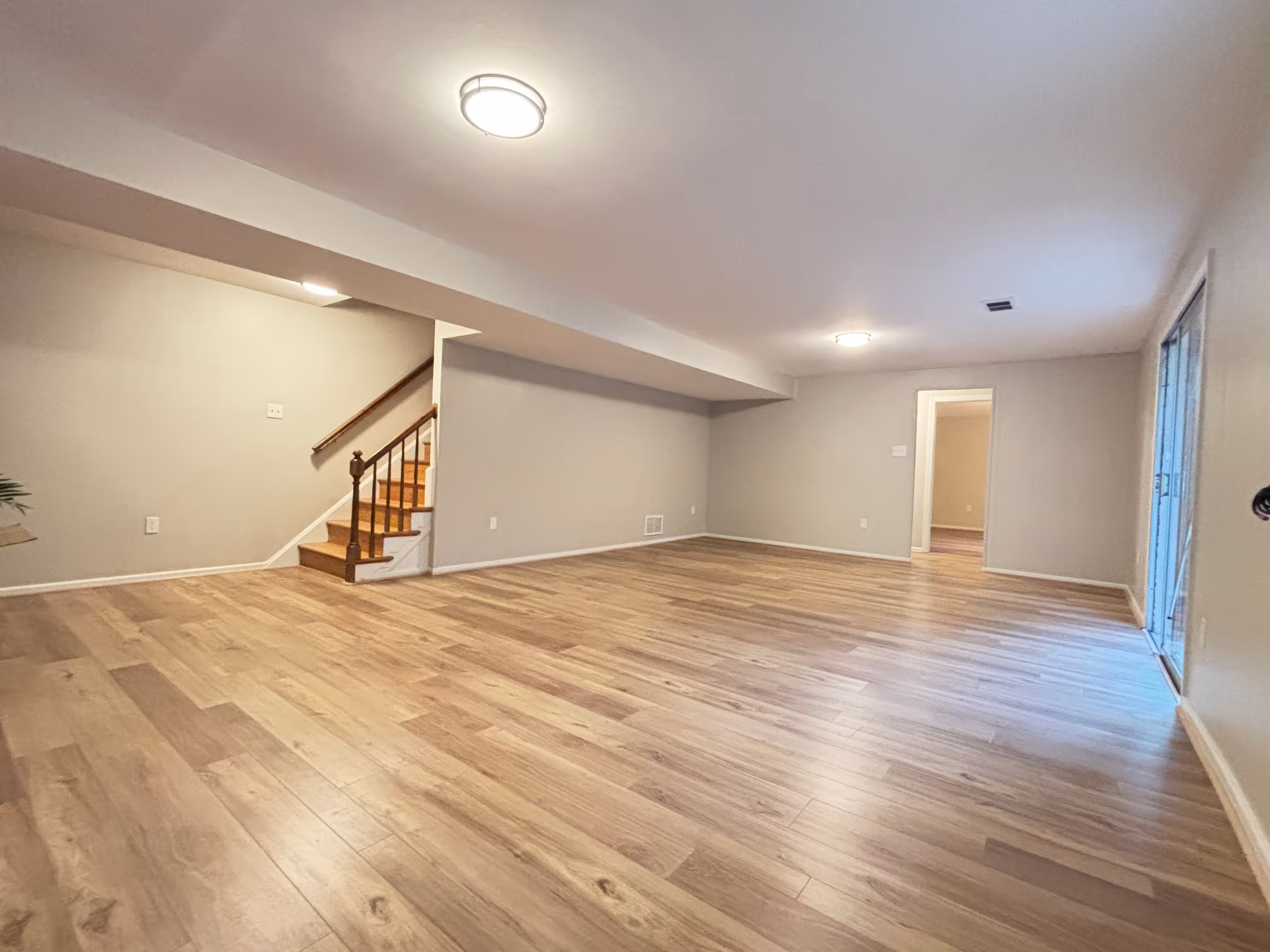 A spacious, newly renovated living area with light gray walls and light-colored wood-look laminate flooring installed throughout. A wooden staircase with white risers and a dark railing is visible on the left, leading to another level. The room is well-lit by a modern flush-mount ceiling light and recessed lighting. A doorway leads to another room with lighter walls, and a sliding glass door on the right opens to the outside. The room is empty and appears clean and modern.