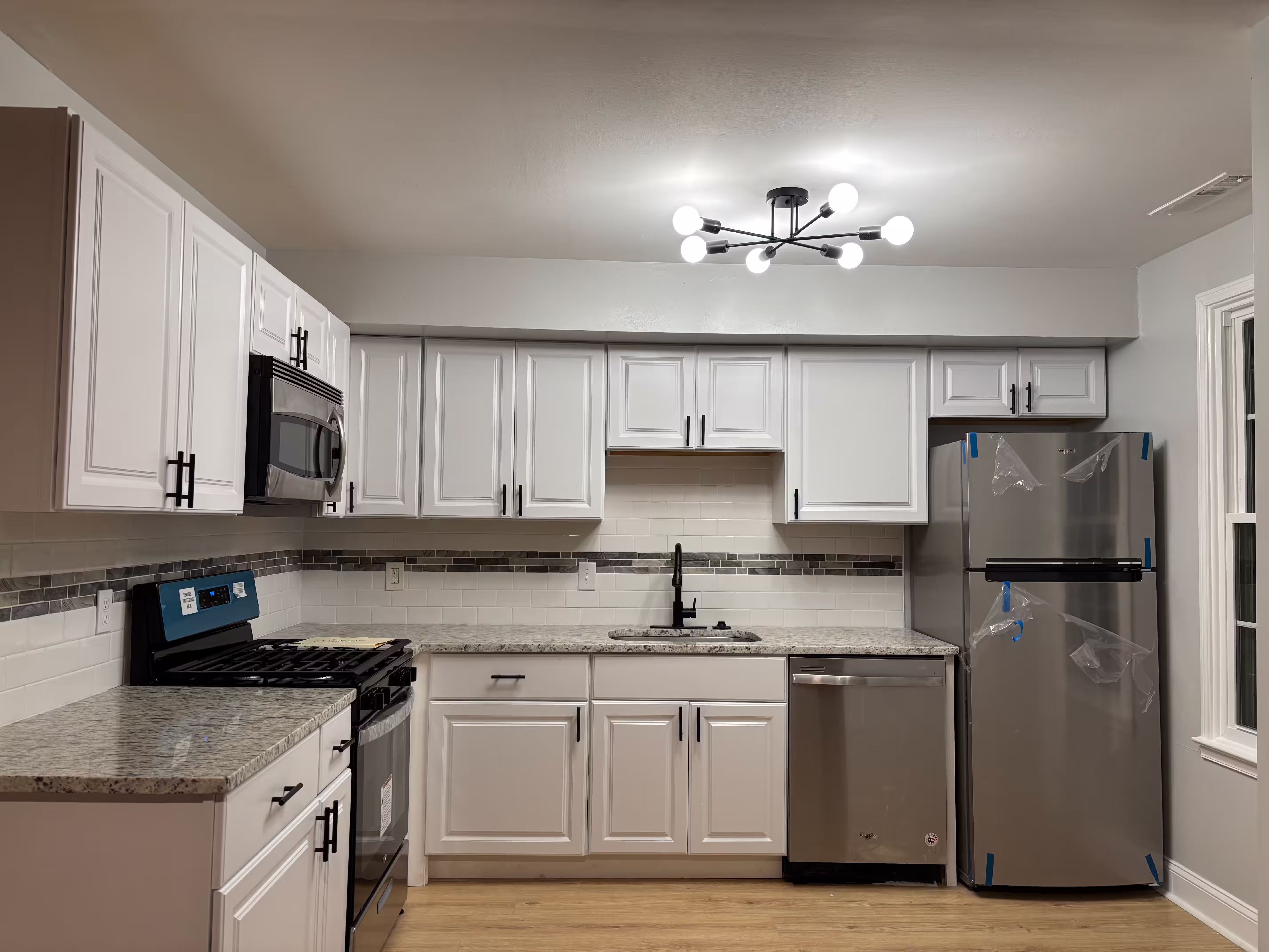 A modern, newly renovated kitchen featuring white shaker-style cabinets with black hardware, light speckled granite countertops, and a light wood-look laminate floor. A stainless steel refrigerator with protective plastic wrap stands on the right. A stainless steel dishwasher is next to a white sink with a black faucet. Above the black gas range is a built-in stainless steel microwave. The backsplash is a combination of white subway tiles and a decorative band of smaller, darker tiles. A contemporary black multi-bulb light fixture illuminates the room from the ceiling.