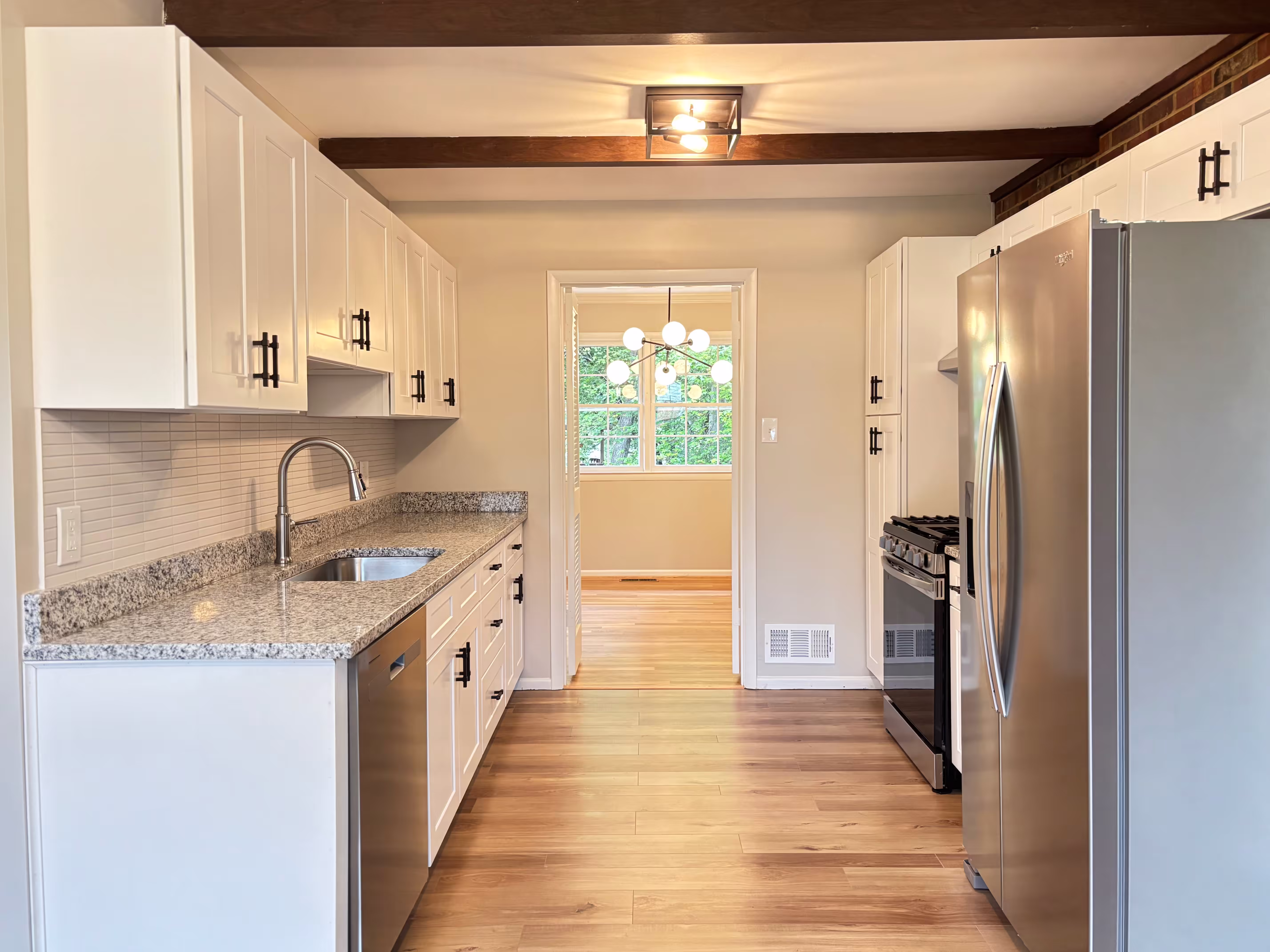 A bright and modern kitchen renovation featuring white shaker cabinets with black T-bar handles, light speckled granite countertops, and a light-toned hardwood floor laid lengthwise. A stainless steel dishwasher is to the left of a stainless steel sink with a modern chrome faucet. A stainless steel refrigerator stands on the right next to a black gas range. The backsplash is made of horizontal white subway tiles. Dark wooden beams run across the ceiling, and a modern geometric light fixture provides illumination. An open doorway leads to a room with light beige walls and hardwood flooring, with natural light streaming in from a window.