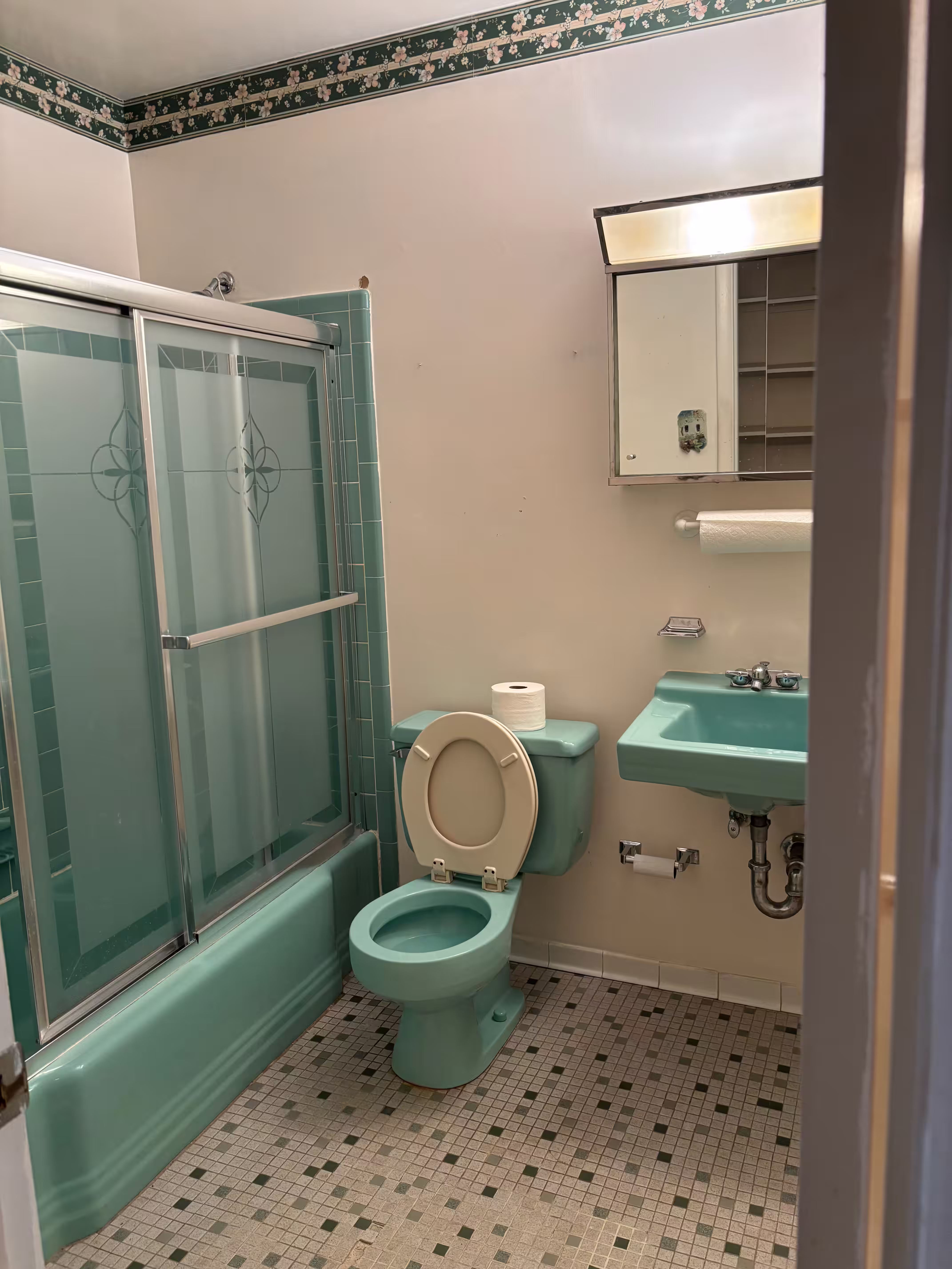 Old bathroom before renovation, featuring vintage mint green fixtures including a toilet, sink, and bathtub with sliding glass doors. The floor has a small tile mosaic pattern in neutral tones, and there's a floral wallpaper border near the ceiling. A mirror cabinet with shelves and a towel holder are mounted on the wall.