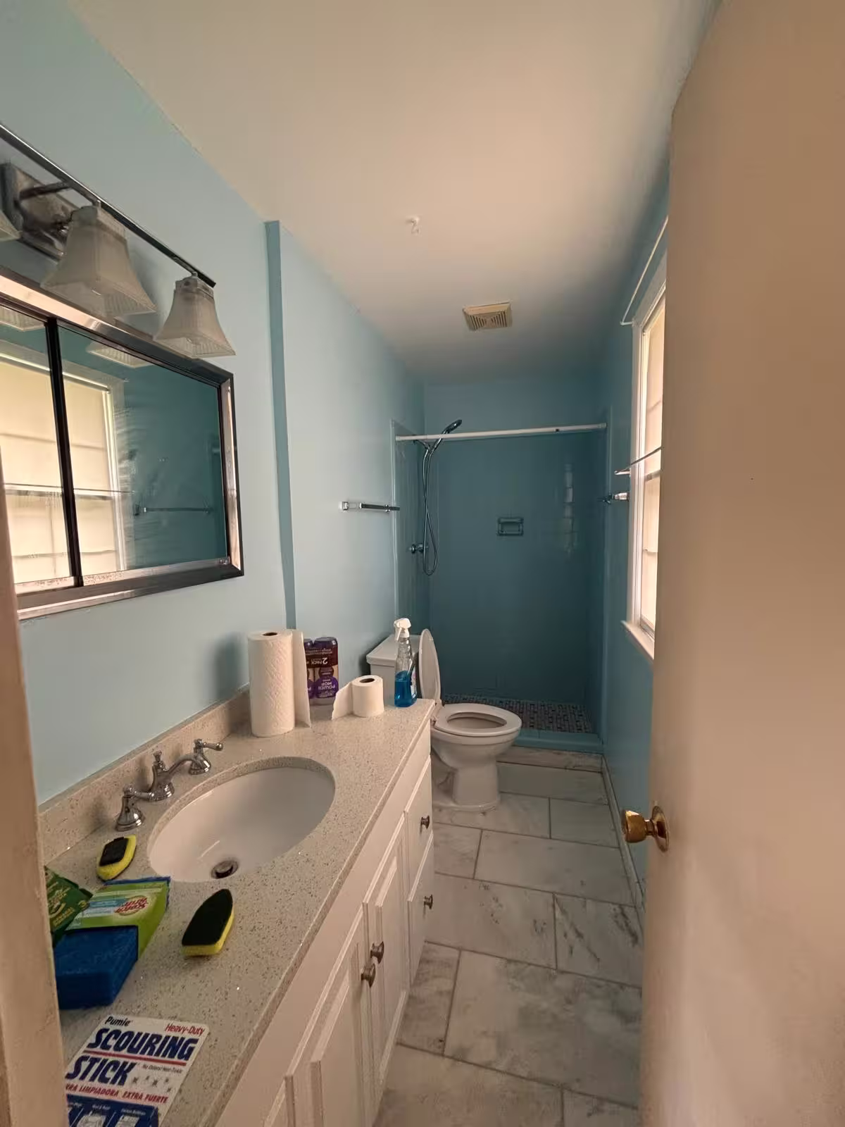 A bathroom with light blue tiled walls in the shower area and the same light blue paint on the remaining walls. The floor is covered in large, light gray square tiles. A white vanity with a light speckled countertop holds a white sink and a chrome faucet. A framed mirror hangs above the sink, flanked by two multi-bulb light fixtures. To the right of the vanity is a white toilet. A partially open white door is visible on the right side of the frame.