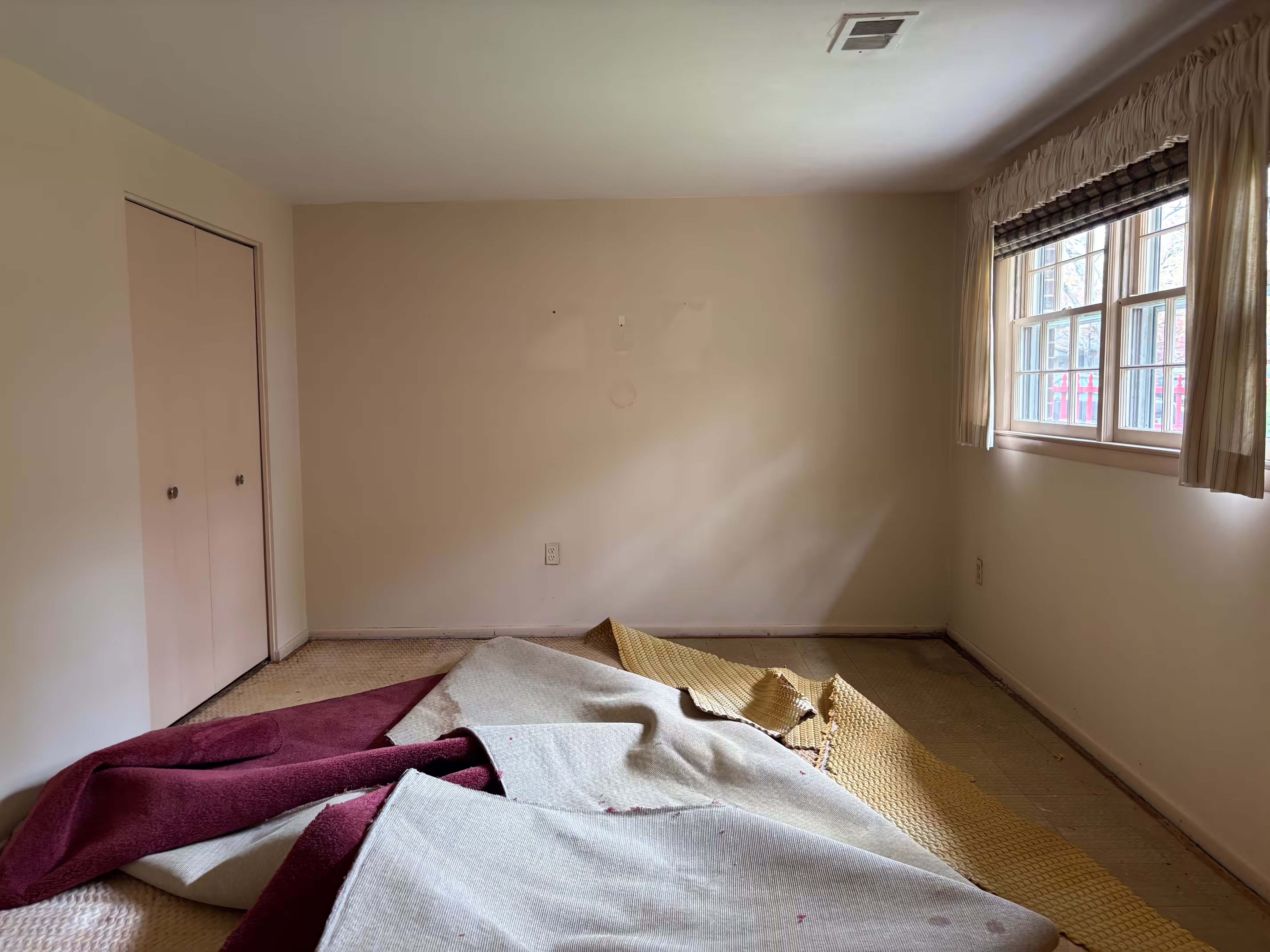 A room undergoing renovation with light beige walls and remnants of old carpet being pulled up. Pieces of maroon, white, and gold carpet are scattered on the floor, revealing a lighter-colored subfloor. A window on the right wall has a brown and white patterned blind and light brown curtains. A closed white double-door closet is visible on the left wall. There are some markings or residue on the wall above where furniture might have been. The room appears to be in the middle of a flooring replacement project.