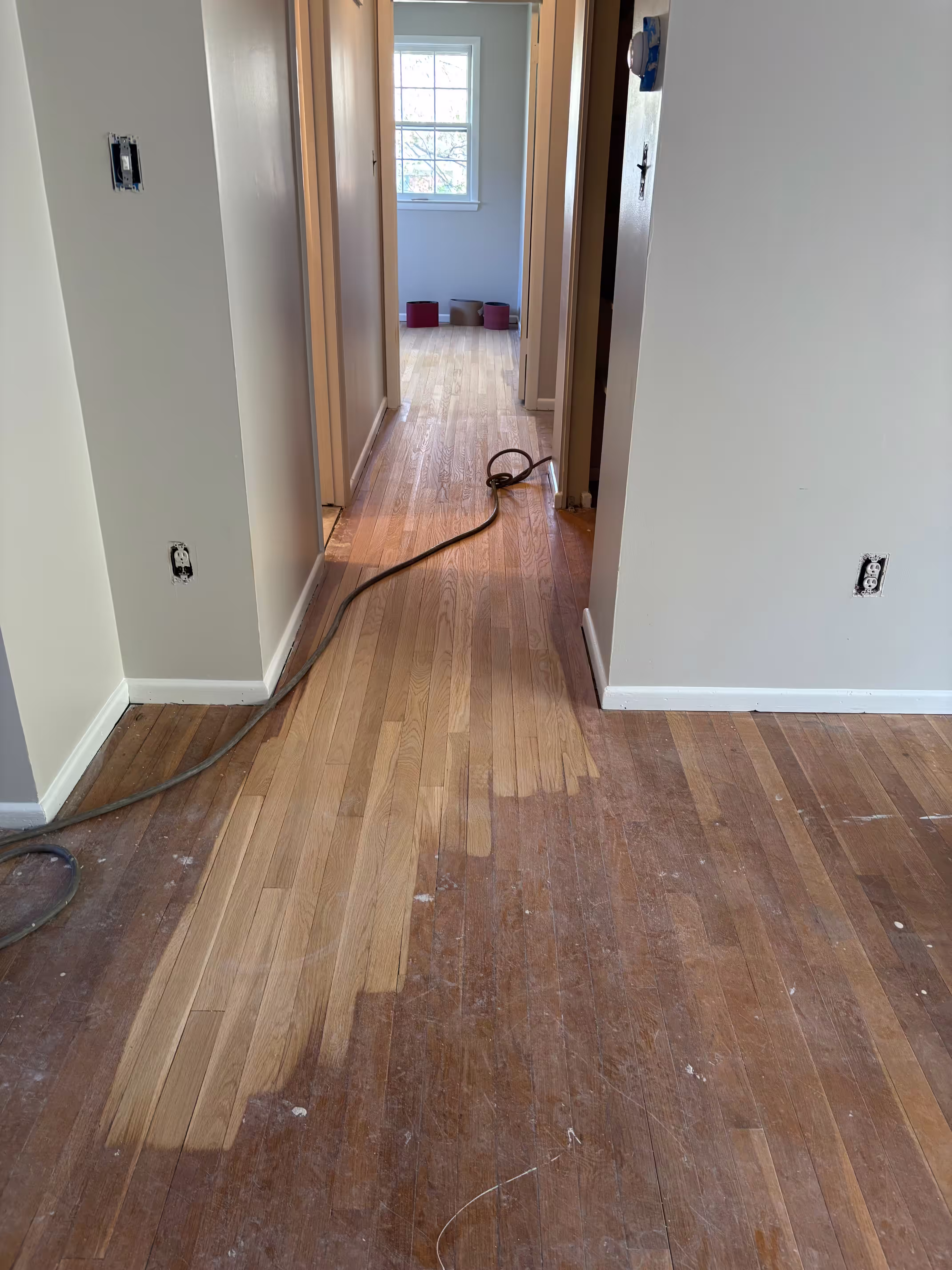 A hallway undergoing floor refinishing. The walls are light beige with white trim. Sections of the original hardwood flooring have been sanded down to a lighter color, while other areas retain a darker, aged finish. Sanding equipment, including a cord and possibly a sander (partially visible on the left), is present on the floor. Electrical outlets on the walls have their cover plates removed. The hallway leads to another room with light blue walls and a window. The contrast in the floor color indicates the sanding process is underway.