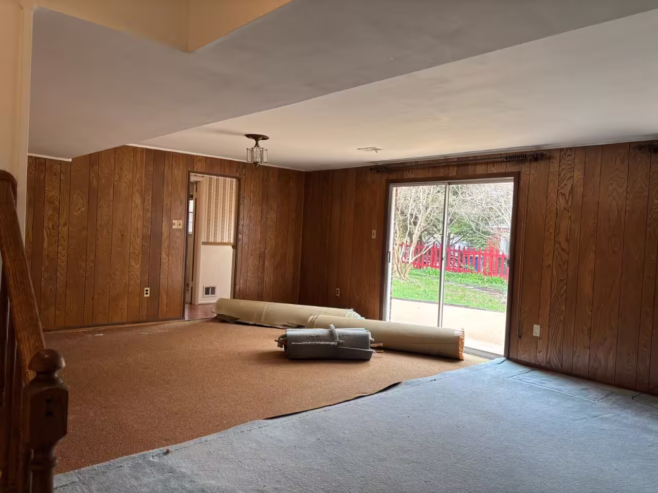 A living room undergoing renovation. The walls are partially covered in dark wood paneling and partially painted a light beige. Old brown carpeting has been partially removed, revealing the subfloor in some areas. Rolled-up sections of the removed carpet are visible in the room. A sliding glass door with a dark frame looks out onto a backyard with a red fence. A doorway with striped wallpaper visible inside leads to another room. The room appears to be in the process of demolition and remodeling.