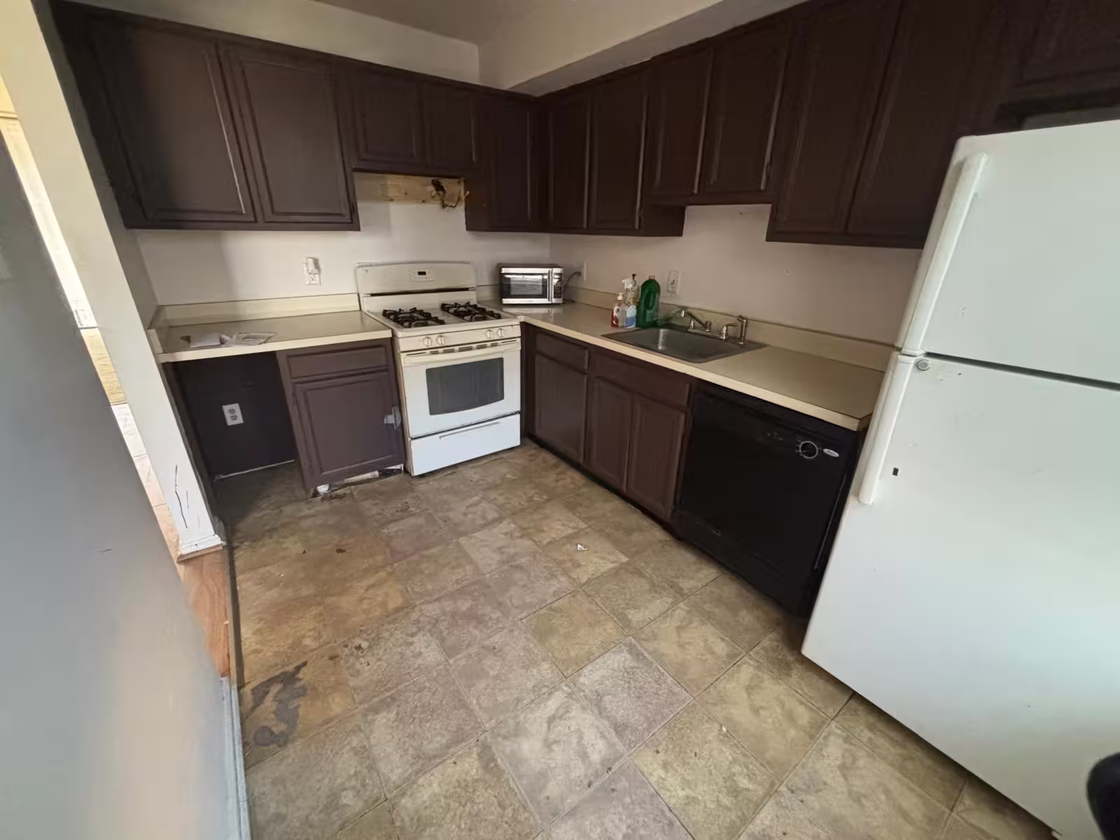 An outdated kitchen with dark brown cabinets, light beige countertops, and a worn, patterned linoleum floor. A white refrigerator is on the right. A black dishwasher is next to a stainless steel sink with a chrome faucet. A white gas range with a black stovetop is against the back wall, with a small toaster oven on the counter beside it. The cabinets show signs of wear and tear, and the overall appearance is dated.
