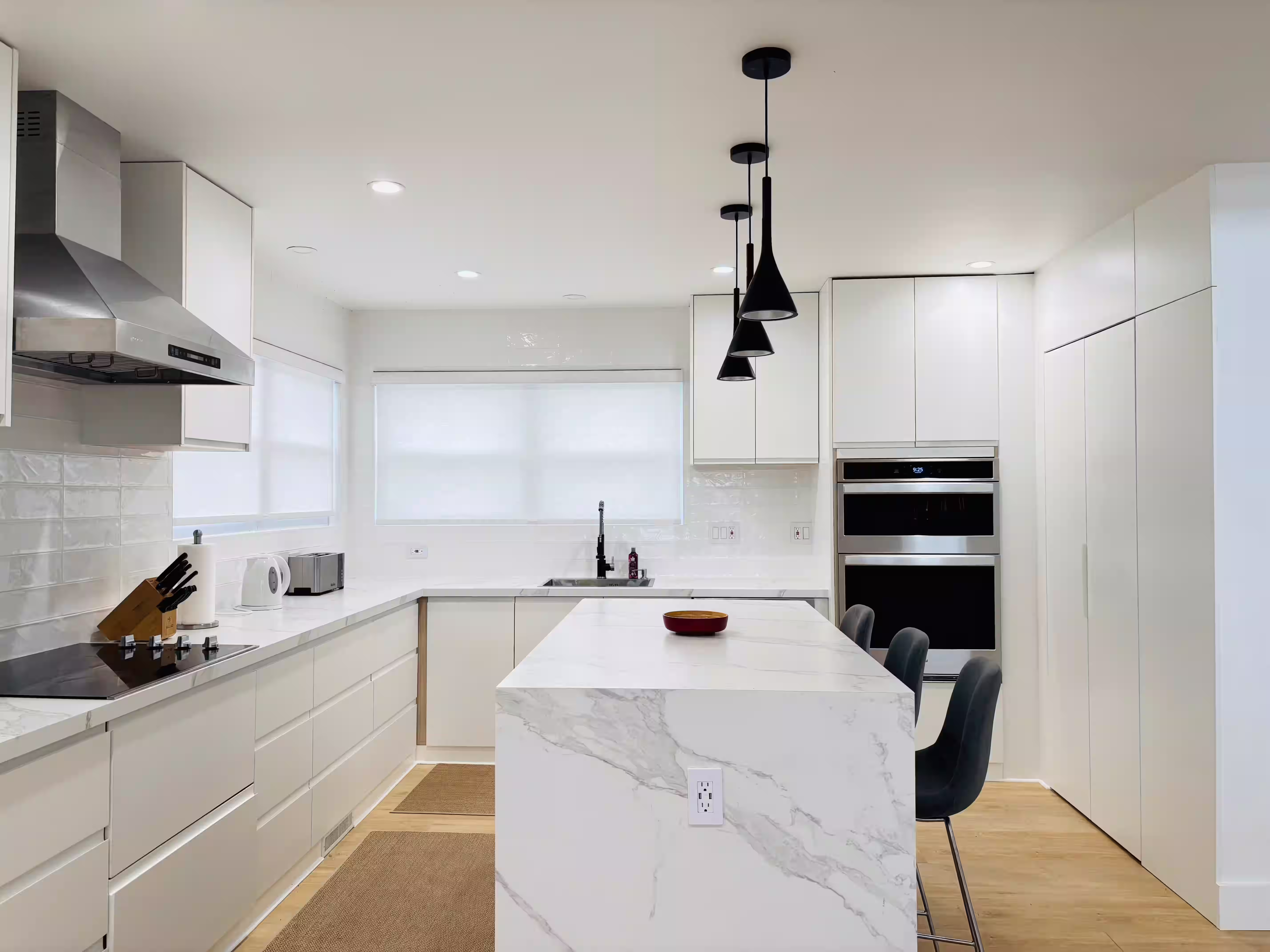 Modern, sleek kitchen with white cabinetry and a large marble island. The space features a built-in cooktop, stainless steel range hood, and pendant lights hanging above the island. A window allows natural light to enter, and the countertops are clean and uncluttered, creating a bright and minimalist atmosphere.