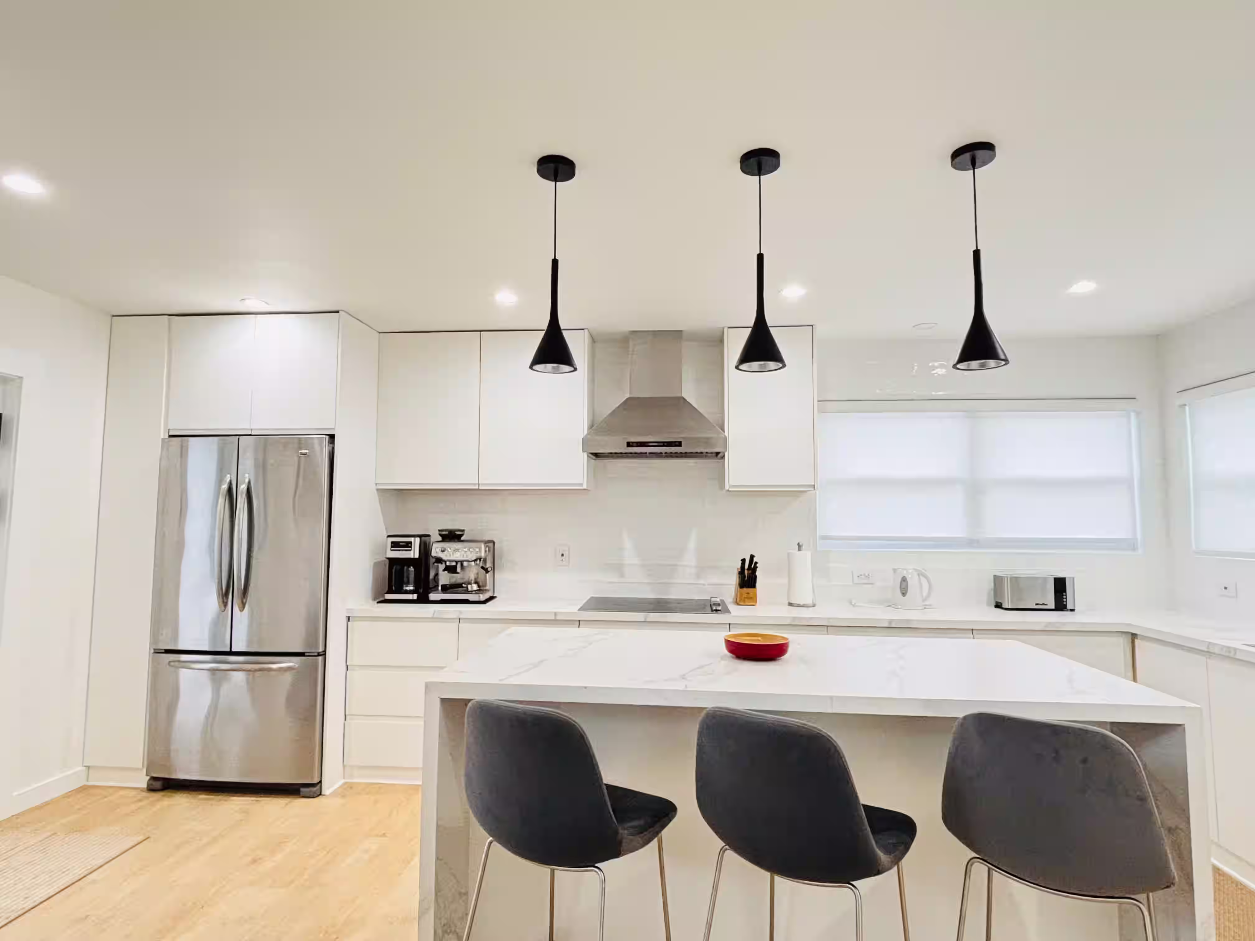 Modern, minimalist kitchen with white cabinetry and a large marble island. The space features sleek stainless steel appliances, including a refrigerator and range hood, along with pendant lights hanging above the island. Three black bar stools are positioned at the island, and the countertops are clean and uncluttered, creating a bright and contemporary atmosphere.