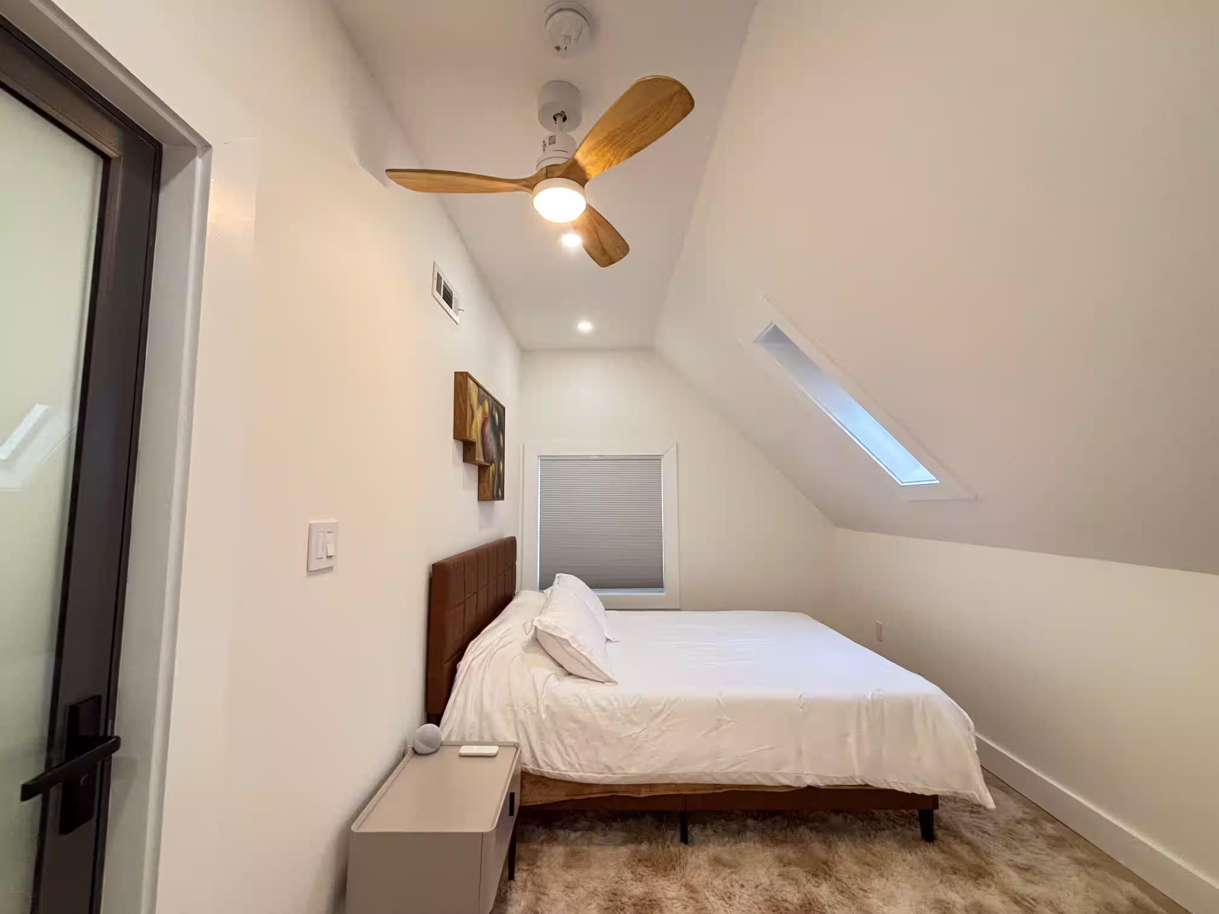 Cozy bedroom with a sloped ceiling and a wooden ceiling fan with a built-in light. The room features a neatly made bed with white bedding and a dark headboard, positioned near a small window with blinds. A skylight allows natural light to enter, and the walls are painted white, creating a bright and airy atmosphere. A minimalistic bedside table is placed next to the bed, and the floor is covered with soft carpeting.