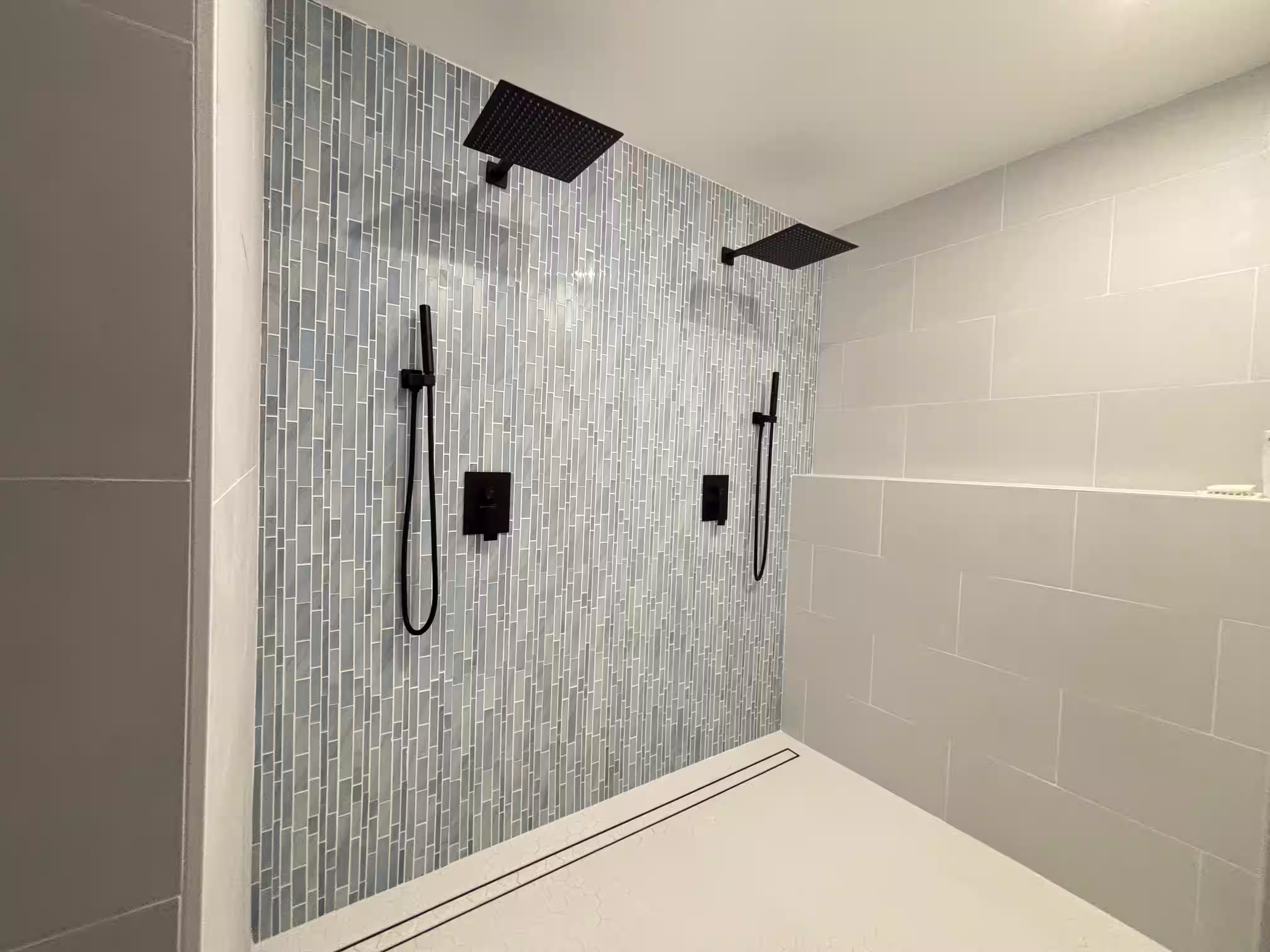 Modern, minimalist shower area with a sleek design. The space features a glass-enclosed shower with a tiled wall in a vertical pattern of gray and blue tones. Two black rain shower heads are mounted on the ceiling, providing a luxurious experience. Additional handheld showerheads are installed on the wall for convenience. The floor is covered with white tiles, and the overall aesthetic is clean, contemporary, and functional.