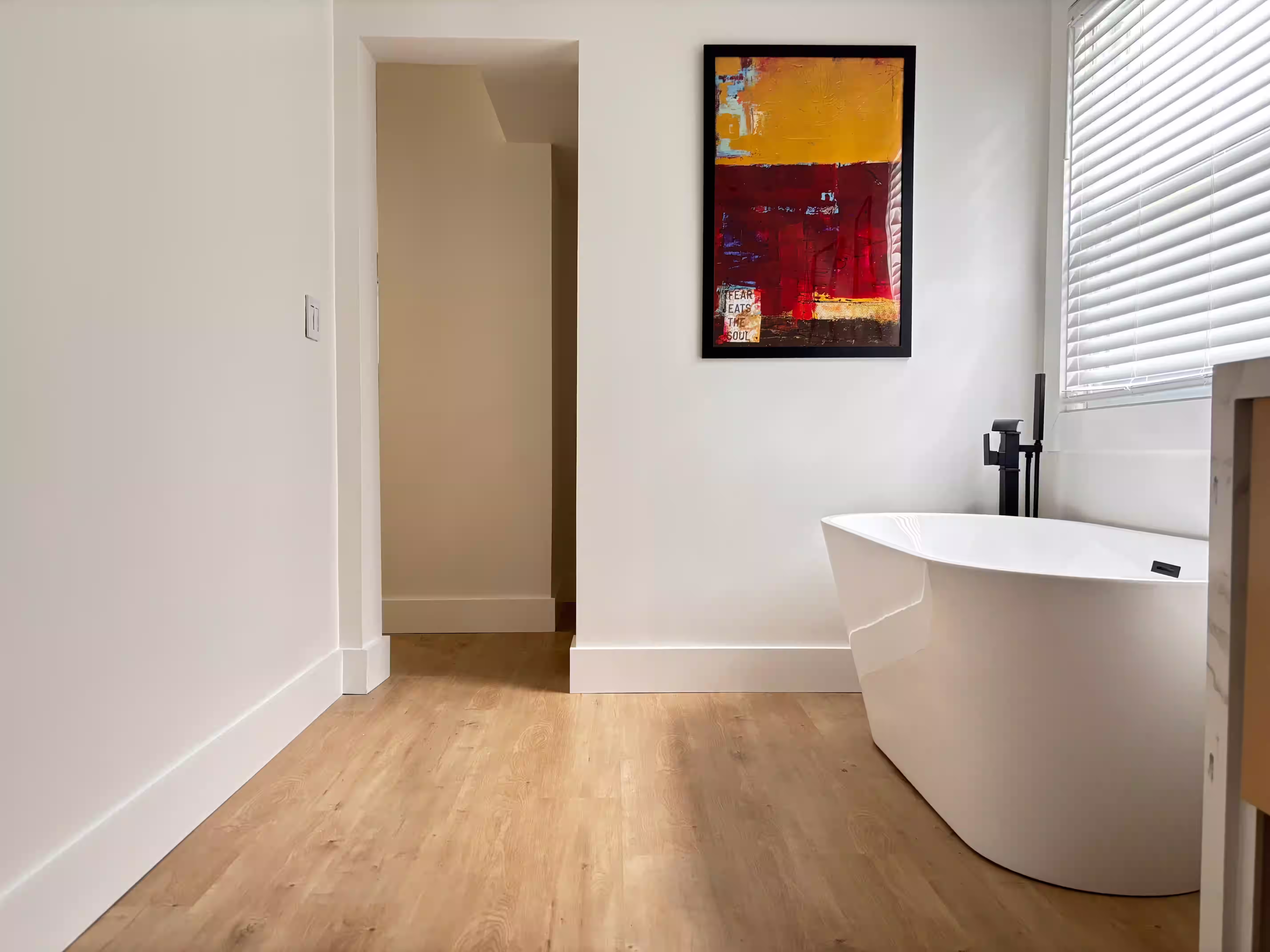 Modern and minimalist bathroom with light wood flooring and white walls. The space features a freestanding bathtub with a sleek, contemporary design and black fixtures. A vibrant abstract painting with bold colors (red, yellow, and black) hangs on the wall above the bathtub, adding a striking visual element. A window with blinds allows natural light to enter, creating a bright and serene atmosphere.