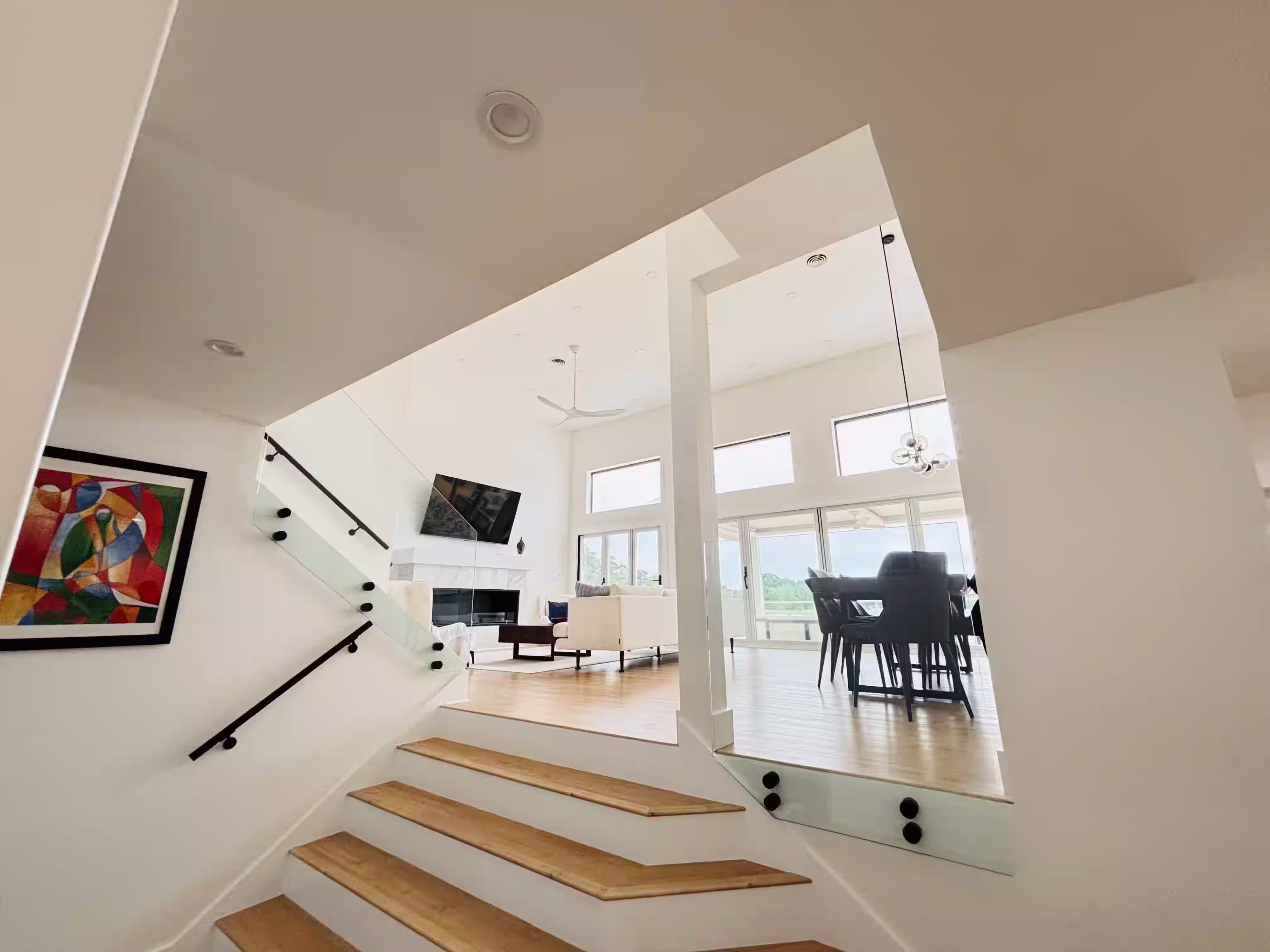 Modern, open-concept staircase with light wood treads and white risers. The railing features a sleek design with black handrails and glass panels, creating a contemporary look. The staircase leads to an upper level that overlooks a bright, airy living area with large windows offering views of the outdoors. A vibrant abstract painting hangs on the wall near the stairs, adding a pop of color. Below, the living space includes a dining area with a table and chairs, and a television mounted on the wall. The overall design is clean, minimalist, and inviting.