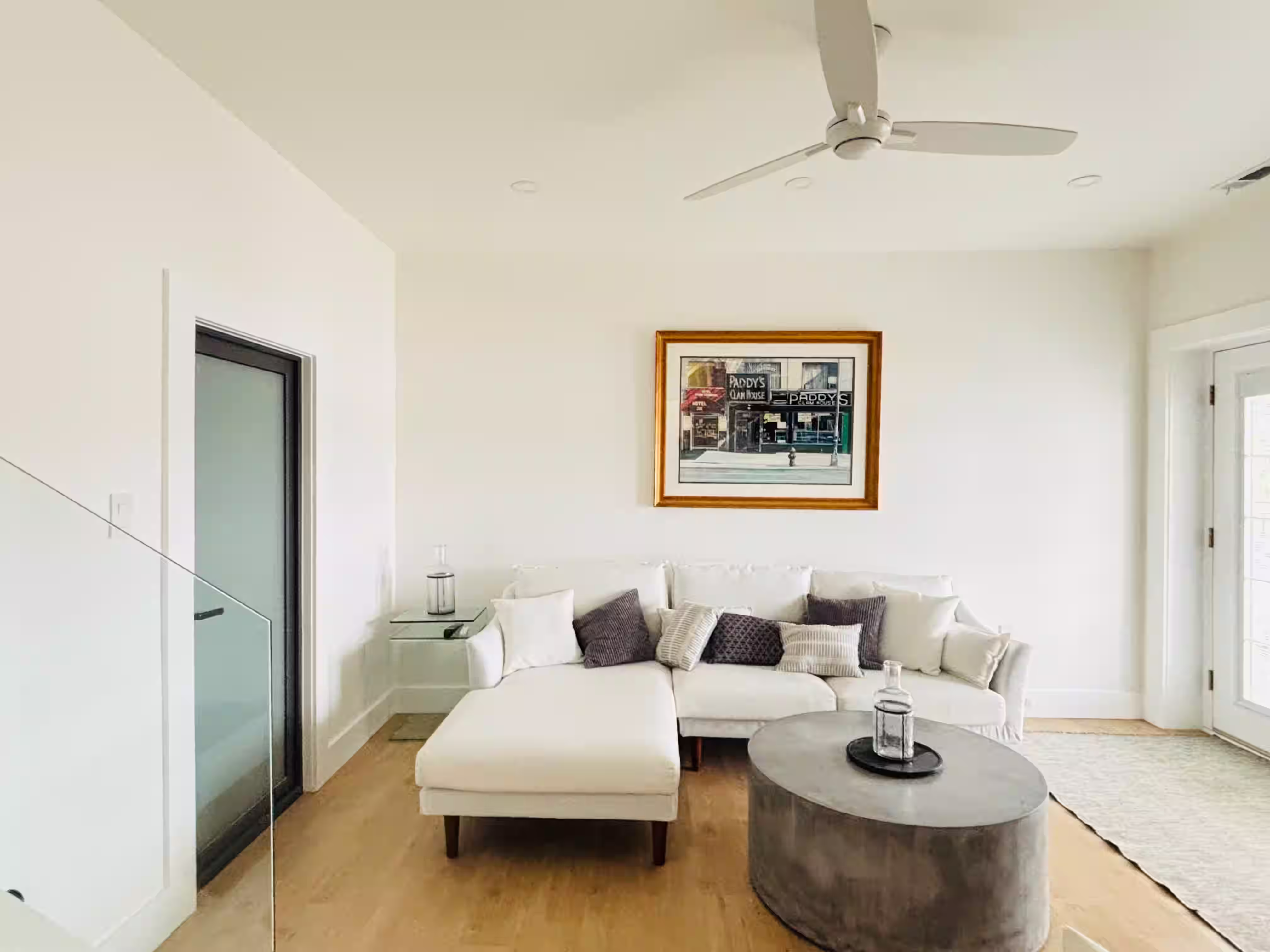 Modern, minimalist living room with light wood flooring and white walls. The space features a sleek sectional sofa in white, adorned with neutral and patterned throw pillows for added comfort and style. A round, textured coffee table sits in the center of the room, holding a bottle on a black tray. Above the sofa hangs a framed artwork depicting a vintage storefront scene, adding a touch of character to the room. A ceiling fan with a simple design provides ventilation, and natural light enters through a glass door on the left side of the room. The overall atmosphere is bright, clean, and inviting.